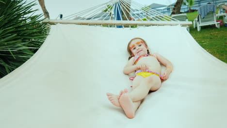 a small girl in a swimsuit and with two pigtails sunbathing on a beach hammock and a resort hotel