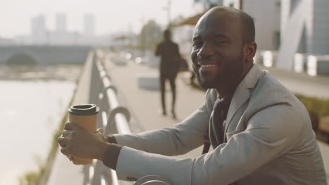 portrait of cheerful african american businessman with coffee on embankment