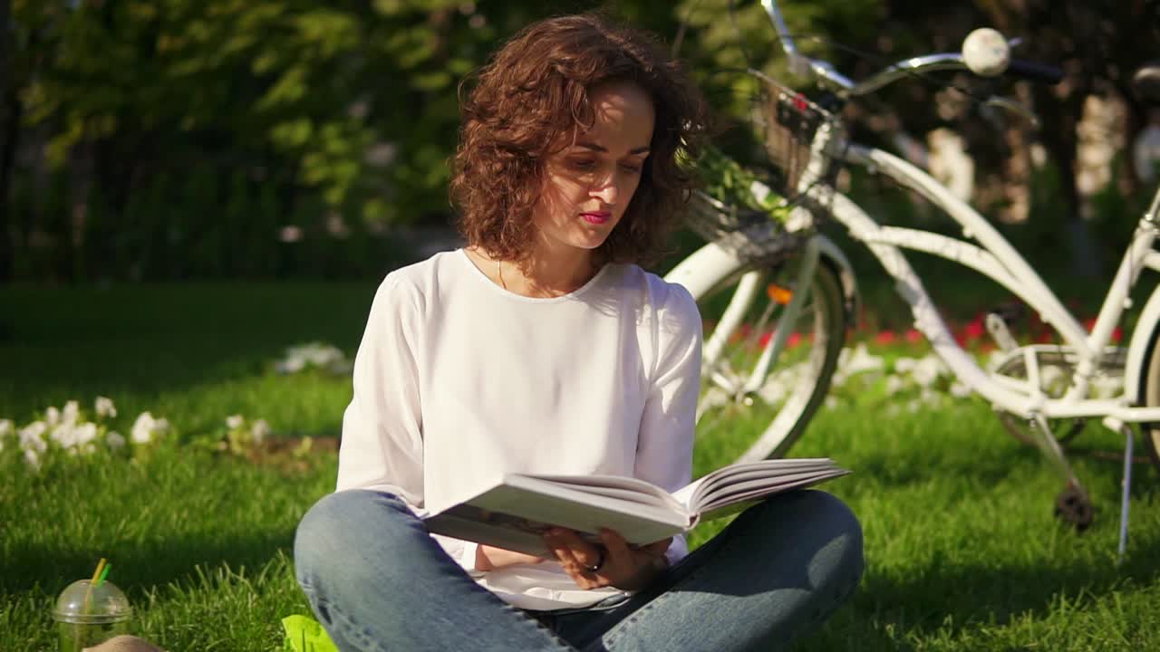 Young woman in white shirt and blue jeans is reading a book sitting on the grass in park and drinking her green detox smoothie
