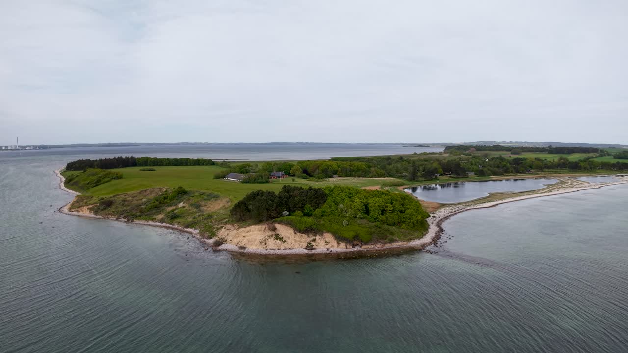 Aerial drone footage of a small peninsula on the Danish coast, showing calm water, sandy shoreline, and lush green vegetation under overcast daylight