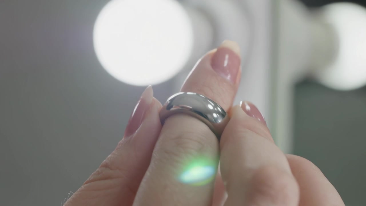 Mani femminili che tengono un anello di metallo, facendolo scivolare e aggiustandolo sul dito medio davanti a una toeletta per farlo calzare, con un bagliore verde.