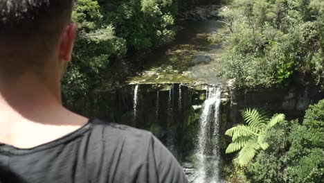 Young-male-tourist-looking-down-at-Mokora-Falls-from-lookout,-Auckland,-New-Zealand