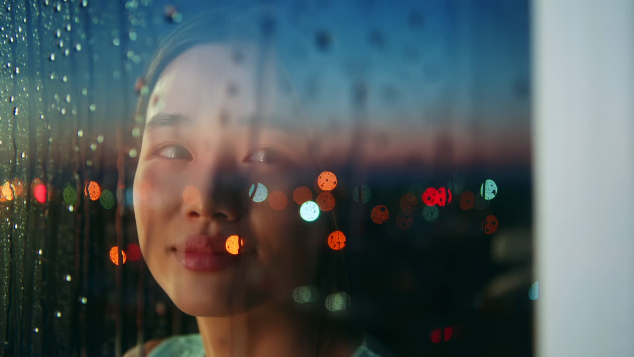 雨粒が散る窓を見つめる若い女性。街の光のボケとガラスの雨滴が、幻想的な反射を生み出している。
