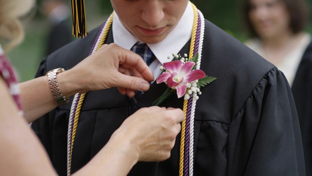 Donna che nota una boutonnière allentata e appunta un'orchidea sulla toga nera di uno studente a una cerimonia di laurea all'aperto.