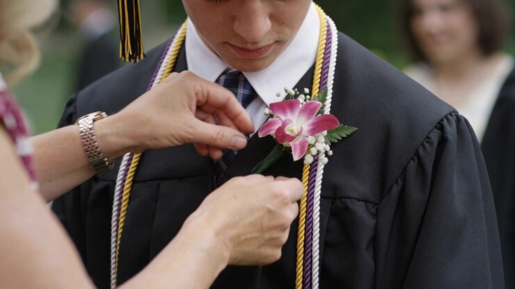 Donna che nota una boutonnière allentata e appunta un'orchidea sulla toga nera di uno studente a una cerimonia di laurea all'aperto.