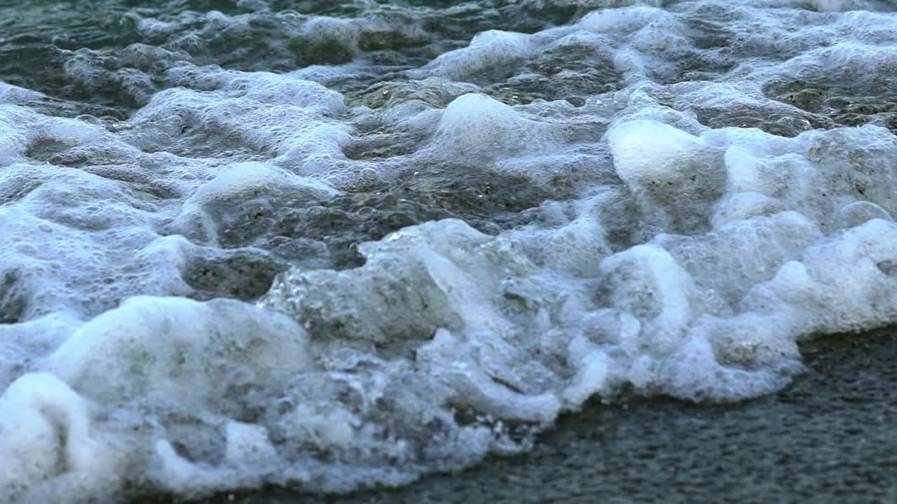 Foamy water background on the coast. Sea waves breaking on the shore and making white foam. Slow motion. Close-up.