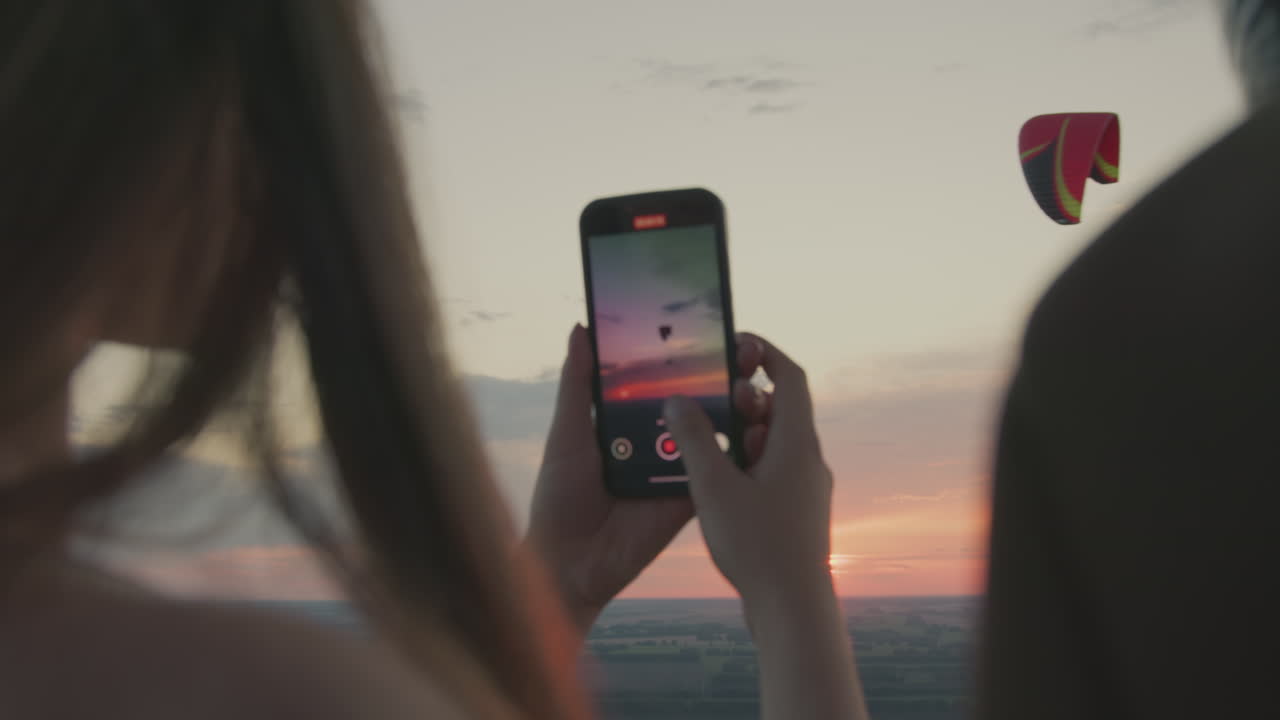 Close rear view of woman holding smartphone to record paraglider soaring across colorful evening sky, with glowing sunset and vast landscape stretching below