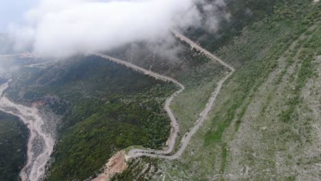 Drone-view-in-Albania-flying-over-a-green-and-rocky-mountain-with-serpent-road-climbing-a-port-on-a-cloudy-day