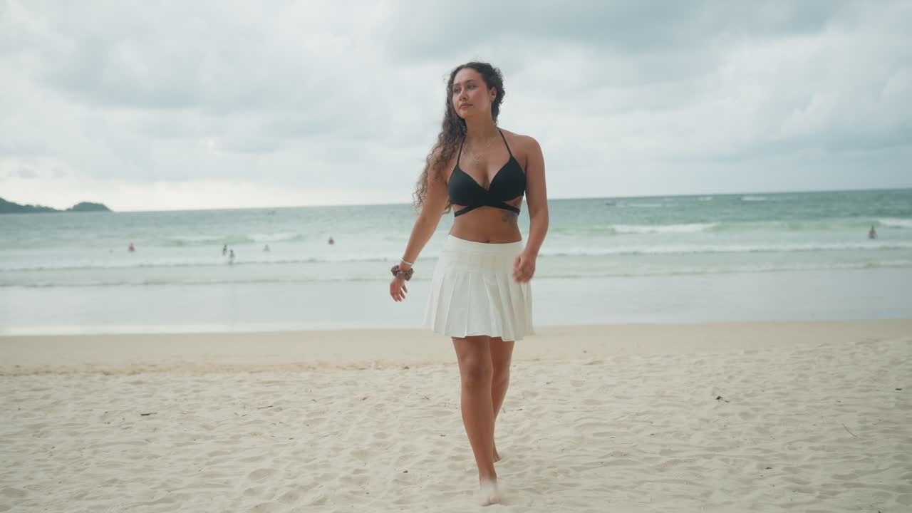 Donna che cammina su una spiaggia isolana sotto un cielo coperto, passeggiata sicura a piedi nudi lungo la riva sabbiosa con onde e isolotti lontani, top bikini nero con gonna bianca, atmosfera di vacanza rilassata e delicata.
