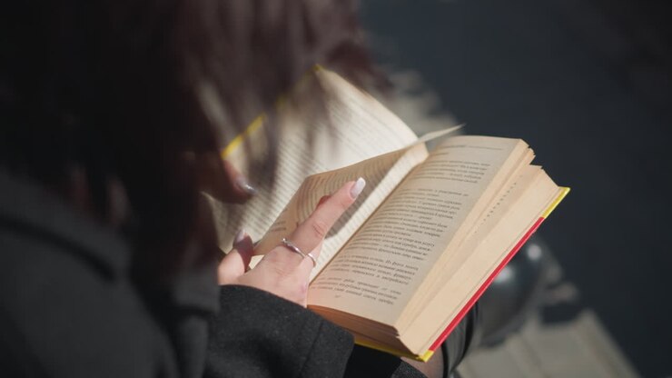 Close-up of a woman reading her book outdoors with hands flipping the page, her nails painted and book with yellow edges is focused in the frame, background features urban setting and lamp posts