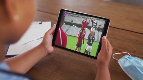 Composite-of-woman-sitting-at-table-with-face-mask,-watching-rugby-match-on-tablet