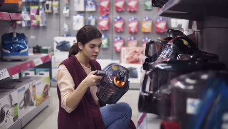 Female-costumer-choosing-new-vacuum-cleaner-in-appliance-store.-Coming-up-to-a-showcase-row-taking-plastic-container-out-and-examining-it-carefuly.-Looking-for-domestic-equipment.