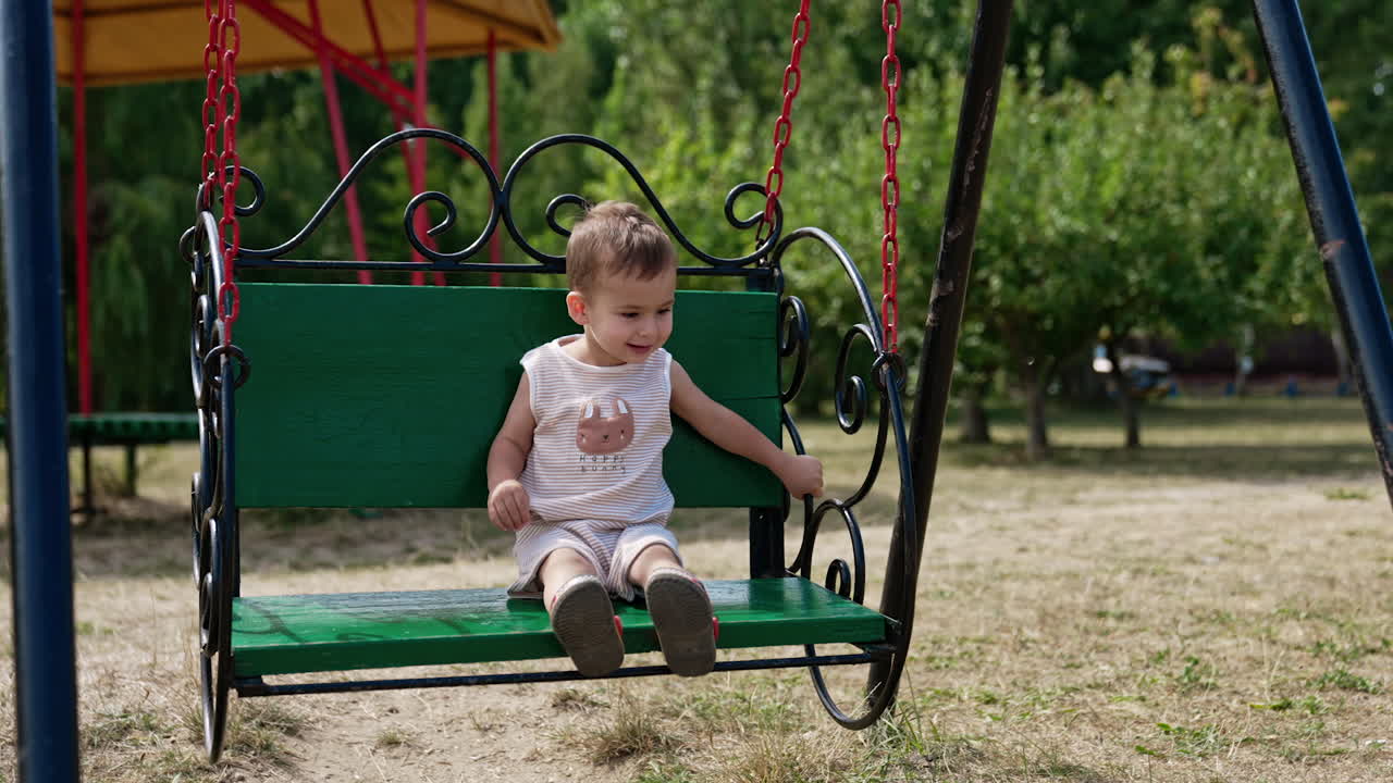 Smiling Caucasian baby boy sits on a swing. Little boy swinging in the park on sunny summer day.