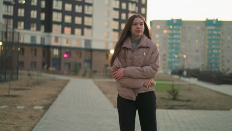 a girl in a peach jacket and black trousers is seen rollerblading through an urban area with tall residential buildings in the background. her expression is dull and pensive, with her hands folded