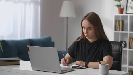 self-education by internet woman is viewing learning video on display of laptop and writing notes in notebook sitting at home young student in living room