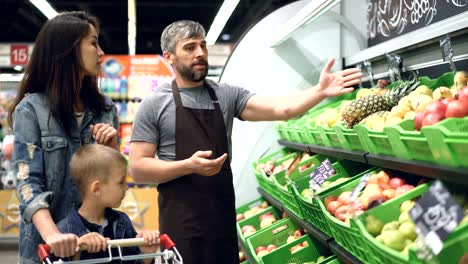 sociable handsome shop assistant is selling fresh fruit to attractive young woman with child, man is pointing at bright boxes with products and speaking.