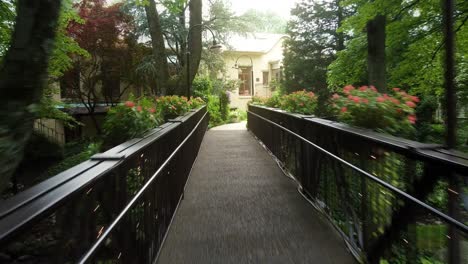 Captura-De-Seguimiento-Rápido-De-Un-Puente-De-Madera-Con-Flores-En-El-Bosque-Que-Conduce-A-Una-Cabaña