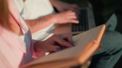 close-up of woman in pink holding book, interacting with her friend typing on laptop nearby, focus on hands and book, blurred background, warm natural light