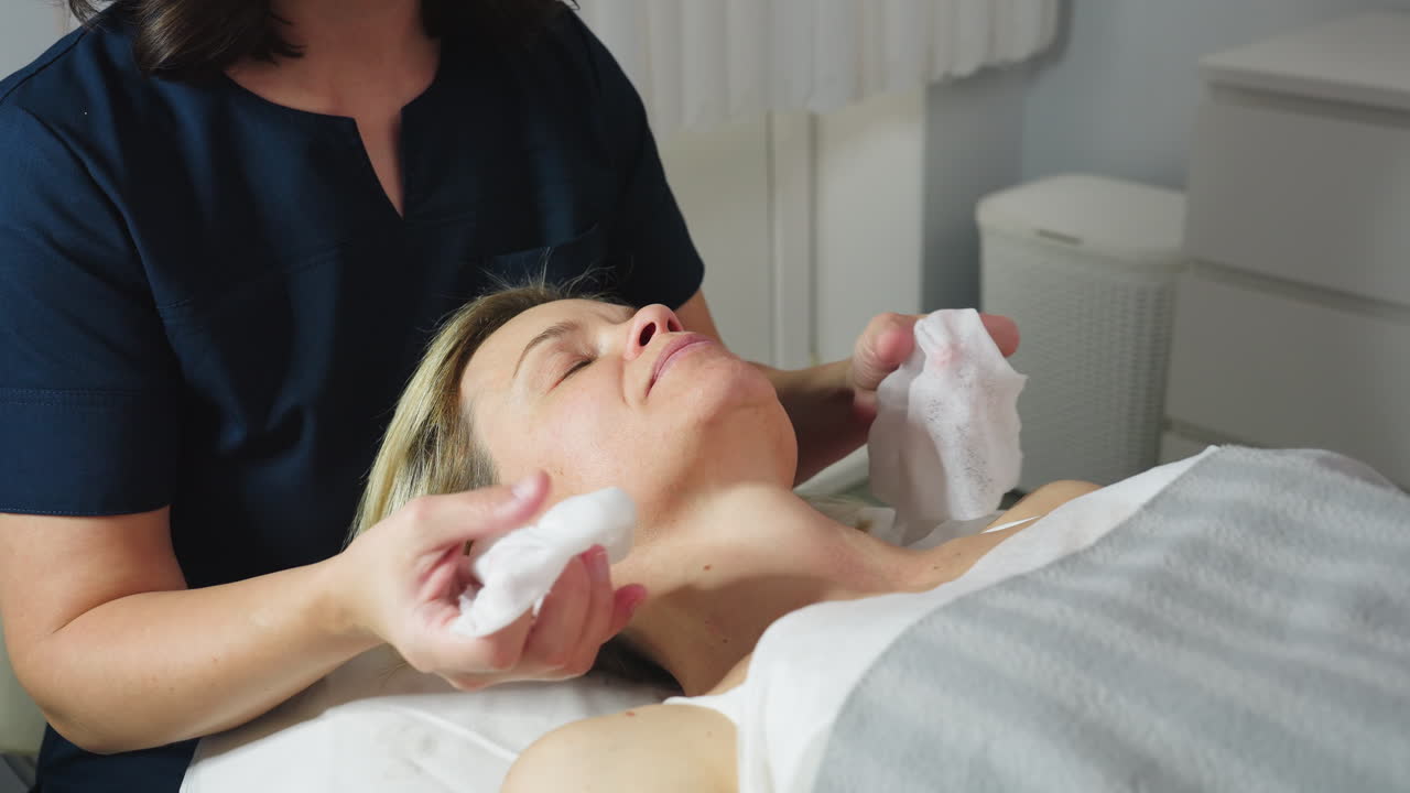 Under bright lamp light massage therapist gently cleanses face of female client with damp cotton pad before skin care treatment in modern white clinic room with calm relaxing atmosphere
