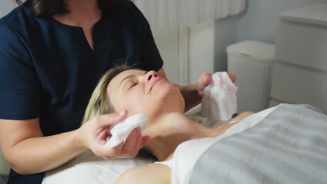 under bright lamp light massage therapist gently cleanses face of female client with damp cotton pad before skin care treatment in modern white clinic room with calm relaxing atmosphere