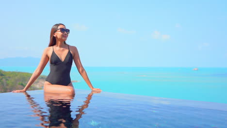 Elegante-Asiatische-Frau-Mit-Schwarzem-Badeanzug-Sitzt-Am-Rand-Des-Infinity-Pools-Mit-Blick-Auf-Das-Meer-In-Zeitlupe