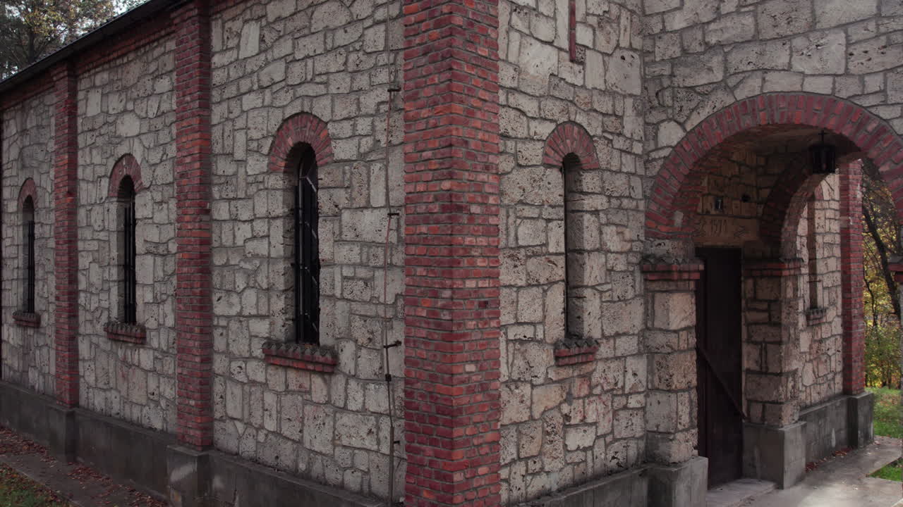 Detailed view of the stone and red-brick entrance of an Orthodox church in Turbe. The historic architecture, arched doorway and window frames stand against a soft autumn forest background