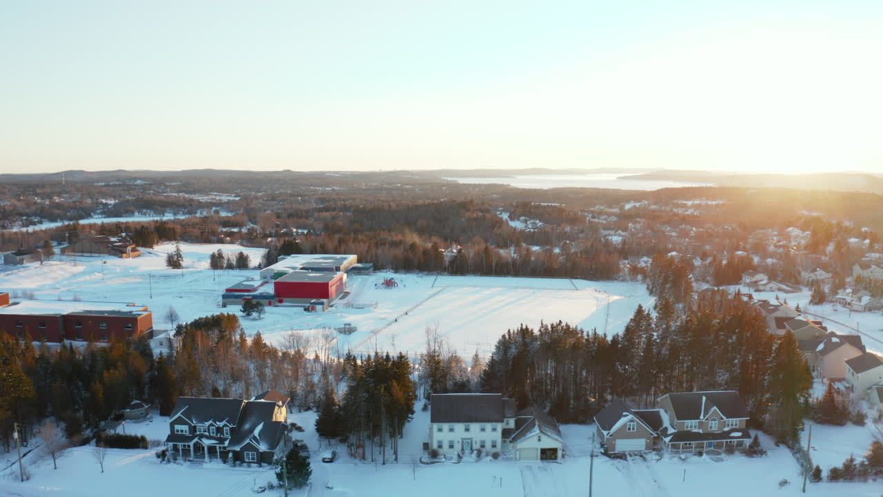 antena escénica de invierno volando sobre casas cubiertas de nieve y un campo escolar al atardecer