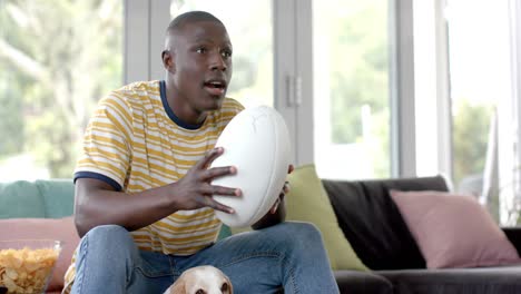 Feliz-Hombre-Afroamericano-En-El-Sofá-Viendo-Un-Partido-De-Rugby-En-Casa,-Con-Su-Perro-Mascota,-Cámara-Lenta