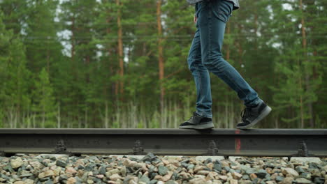 a close-up view of a person wearing blue jeans and black sneakers walking along a railway track, balancing carefully, the background features lush green trees