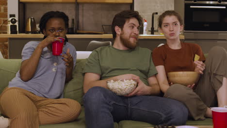 tres compañeros de cuarto están viendo una película mientras comen palomitas de maíz y papas fritas sentados en el sofá en casa 2