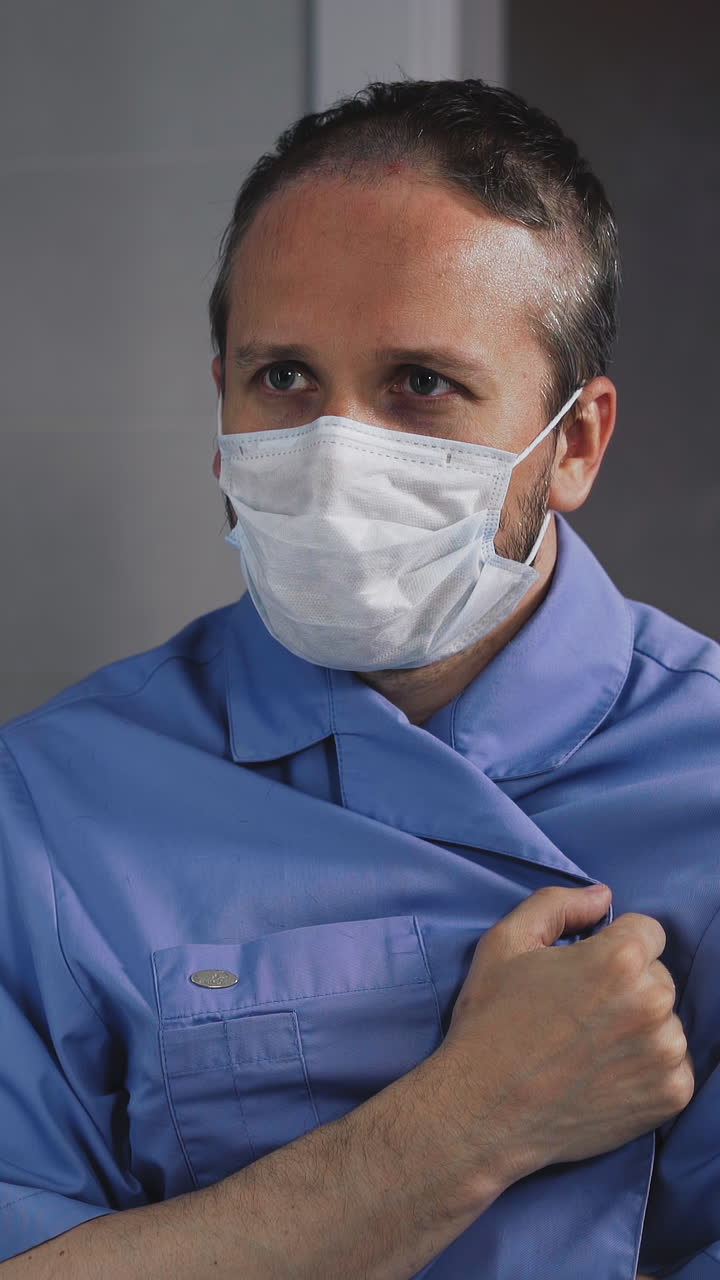 closeup portrait of young uncut doctor, in surgical suit, in front of mirror, in protective mask, unbuttons clothes, looks at his bare chest, then shamefully smells, looks around