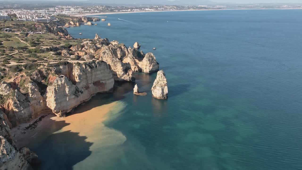 Aerial drone smoothly pans up from dramatic limestone cliffs and stacks, unveiling the sweeping coast, turquoise waters, and sandy beaches stretching toward Lagos in Portugal’s Algarve
