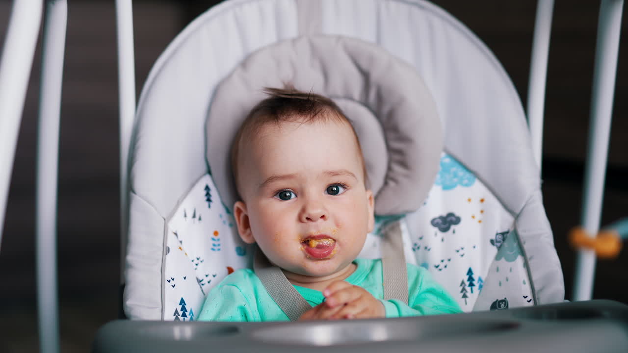 Baby boy nutrition at mealtime. Adorable baby is given food and he waves hands cheerfully and shows tongue.