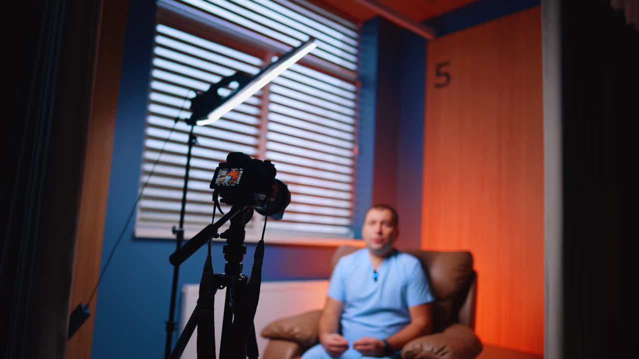 Behind Scenes Doctor Vlog Recording In Clinic Room. Behind scenes view of camera filming doctor in clinic room with orange wall and window blinds