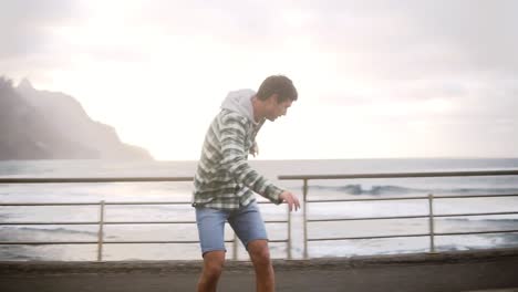 sport, lifestyle, extreme and people concept - tall guy riding longboard by the coastline road in cloudy weather. full length of an active man exercising on road. slow motion. cloudy weather