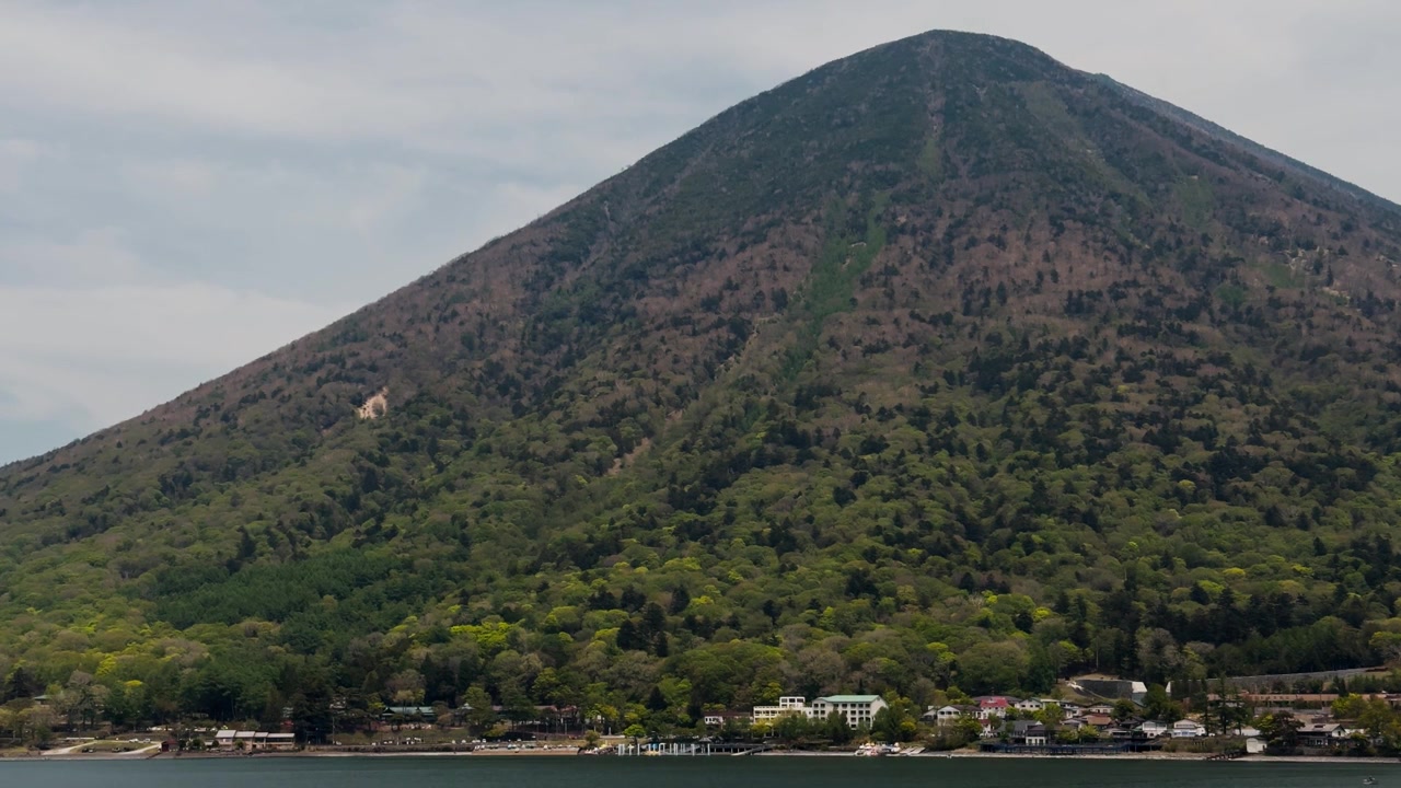 Malerische Aussicht auf einen steilen bewaldeten Vulkanberg über einem friedlichen Dorf am See und ruhigem Wasser in der japanischen Landschaft