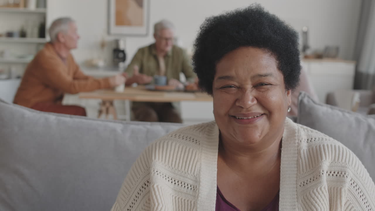 Chest-up POV of happy middle-aged African woman sitting on couch, looking up and smiling on camera