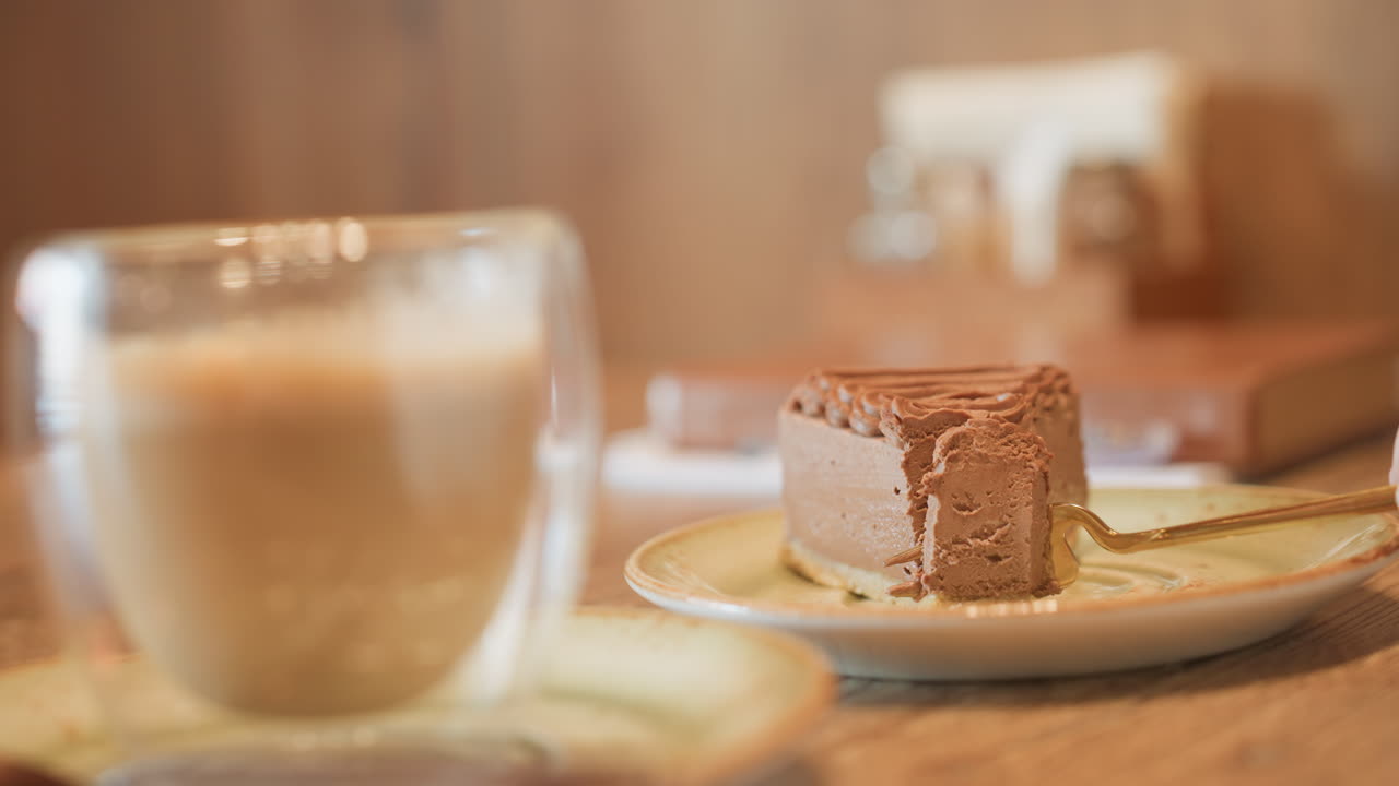 close up glass of milkshake in foreground with customer using fork to slice chocolate cake on wooden table, background softly blurred, cozy cafe atmosphere