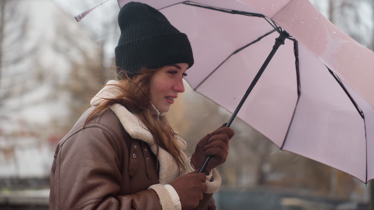 Close shot of young girl wearing black knit cap and brown shearling jacket holding umbrella as wind blows gently with light snowfall, strands of hair in motion, showcasing winter chill and calm