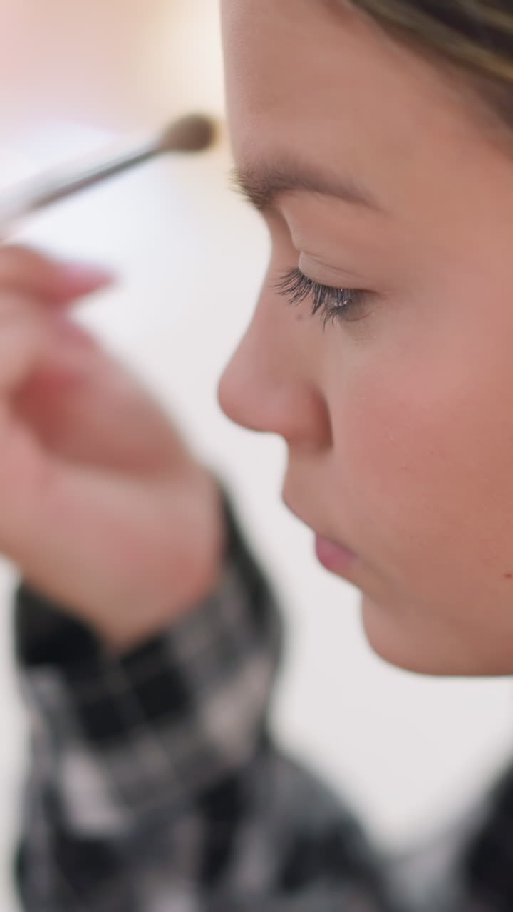 브러시, 격자무늬 셔츠, 부드러운 햇빛, 클로즈업 프로필로 내추럴 메이크업을 마무리하는 십 대 소녀. 눈썹과 뺨을 가로지르는 부드러운 터치, 편안한 표정, 은은한 마스카라, 깔끔하고 미니멀한 팔레트