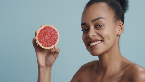 Porträt-Einer-Attraktiven-Jungen-Afroamerikanerin-Mit-Grapefruit,-Die-Lächelnd-Die-Natürliche,-Gesunde-Hautpflegeessenz-Genießt.-Schöne-Frau-Mit-Perfektem-Teint-Auf-Blauem-Hintergrund