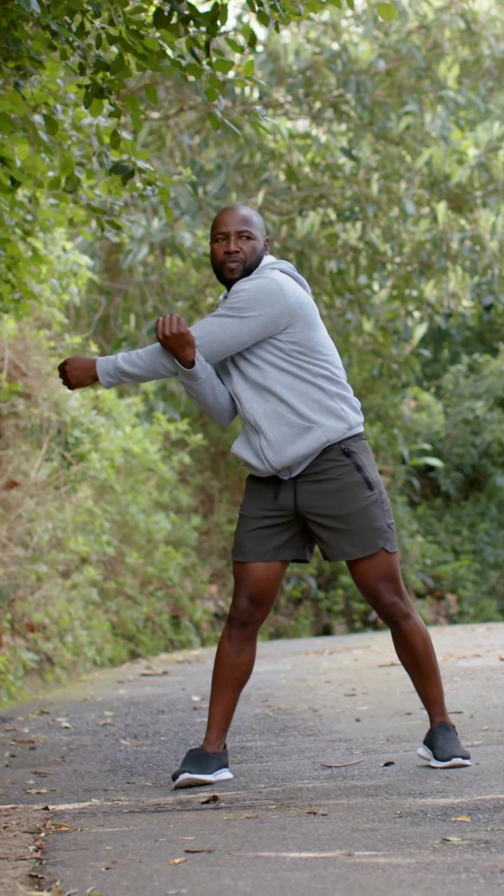 Vertical video: Doing crossbody stretch, man in gray hoodie doing torso twists warming up on path