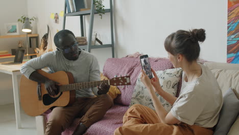 mujer filmando a su marido con el teléfono mientras toca la guitarra en casa