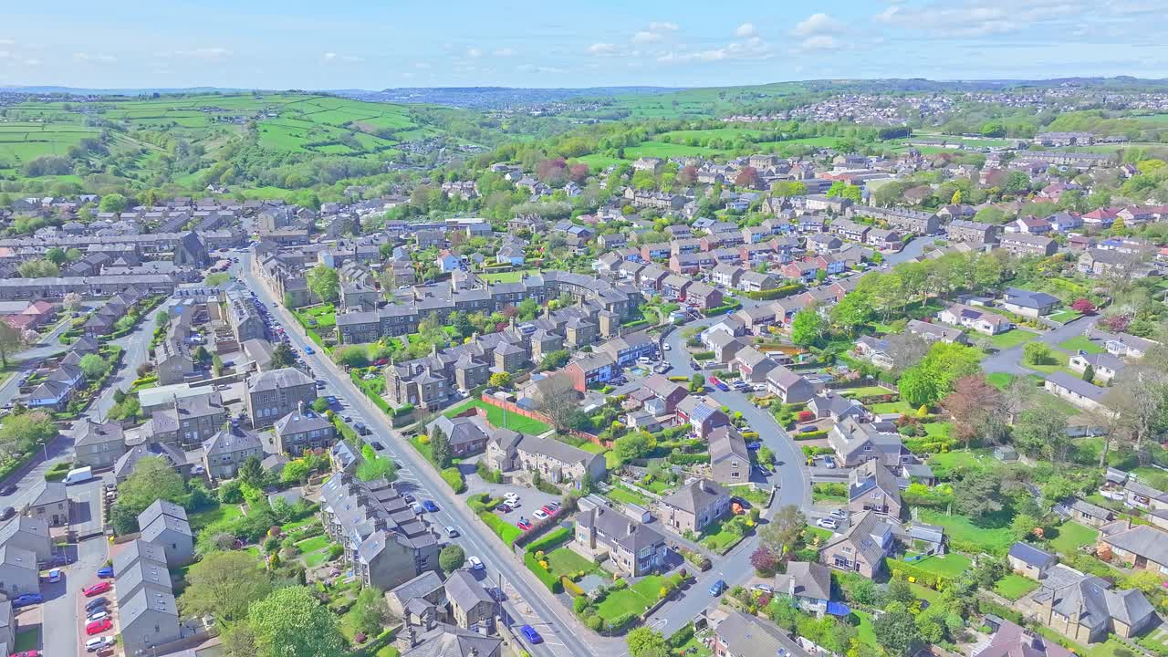 Stationary aerial capture of Shibden Park in Halifax, West Yorkshire, showing blooming cherry trees, open lawns, and the surrounding residential area beneath a clear sky, slow motion