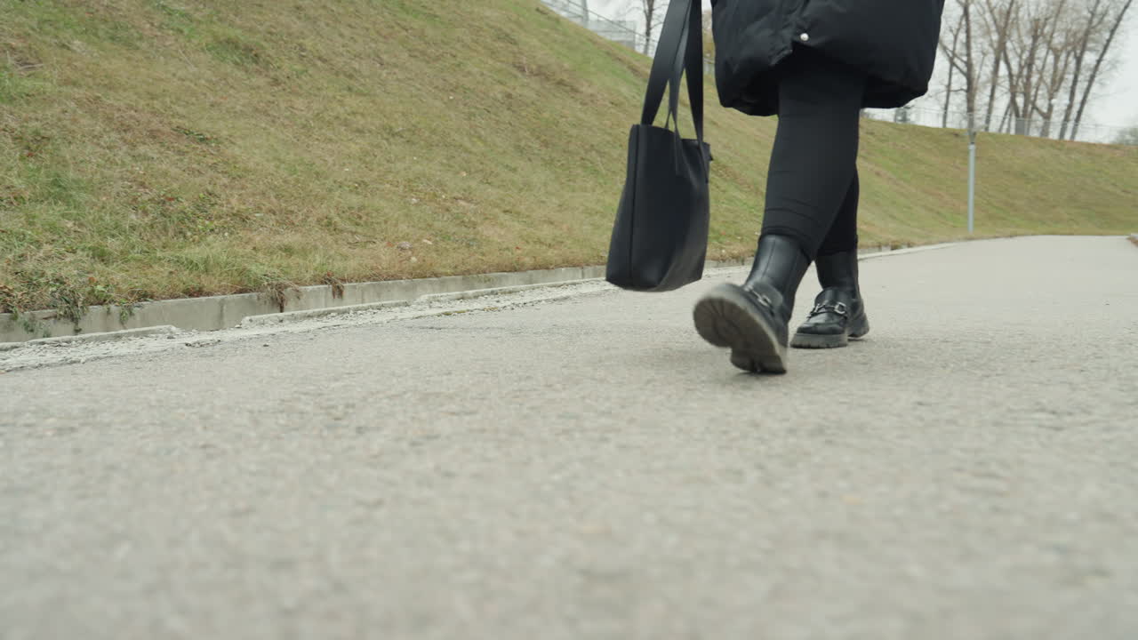 Primer plano de ángulo bajo de pavimento, mujer caminando con abrigo de invierno negro llevando un bolso tote, pasos lentos y firmes en un camino suburbano nublado, botas y cordones visibles, bordillo y borde de césped, tonos apagados.