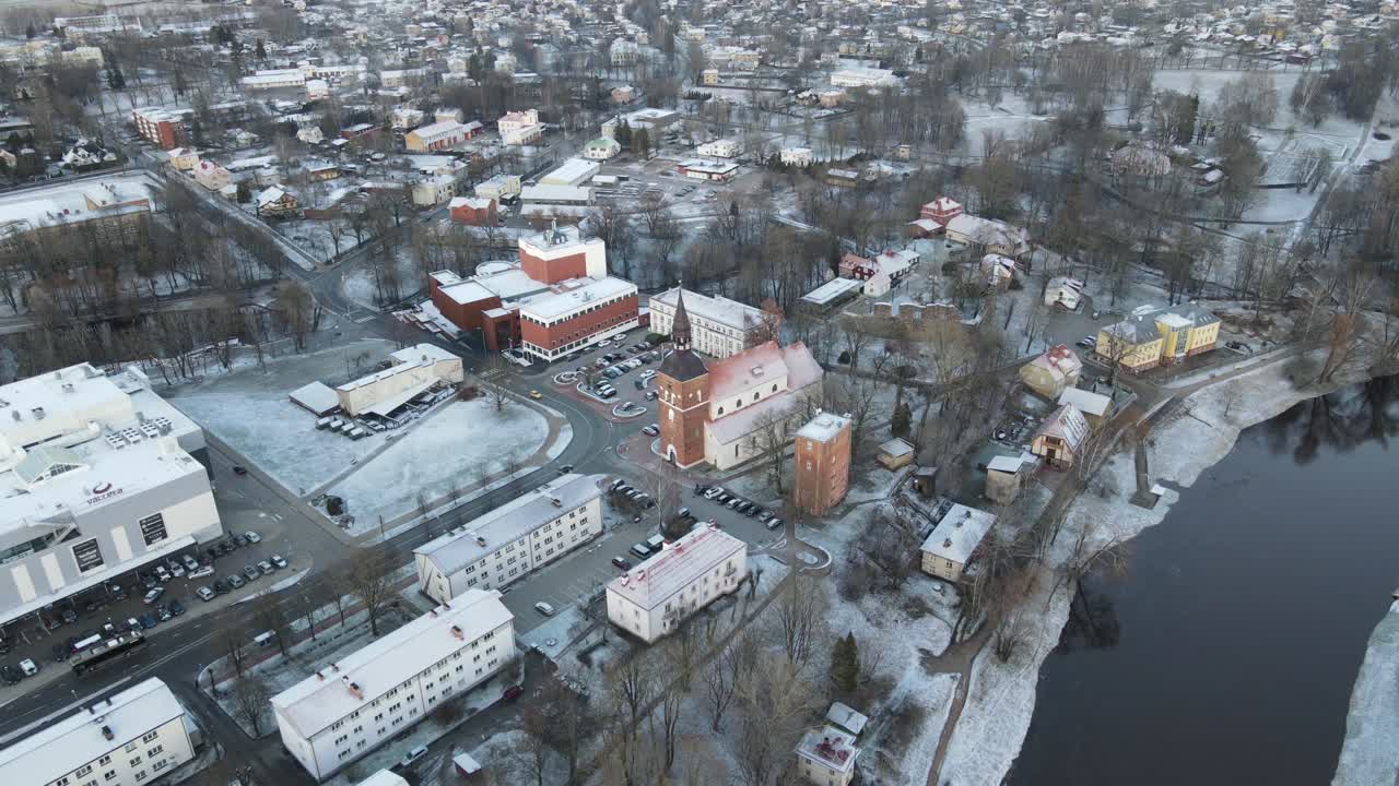 A breathtaking aerial view of Valmiera city, showcasing the winding Gauja River surrounded by snow-covered landscapes and residential areas under a wintry sky.