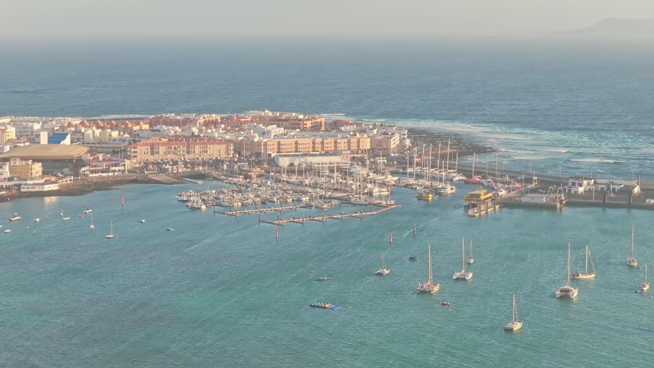 Drone ascend over Playa del Medio Beach Area at sunrise in Fuerteventura, Canary Islands, Spain, revealing a coastal marina, sunken vessel, and surrounding townscape bathed in golden Atlantic light