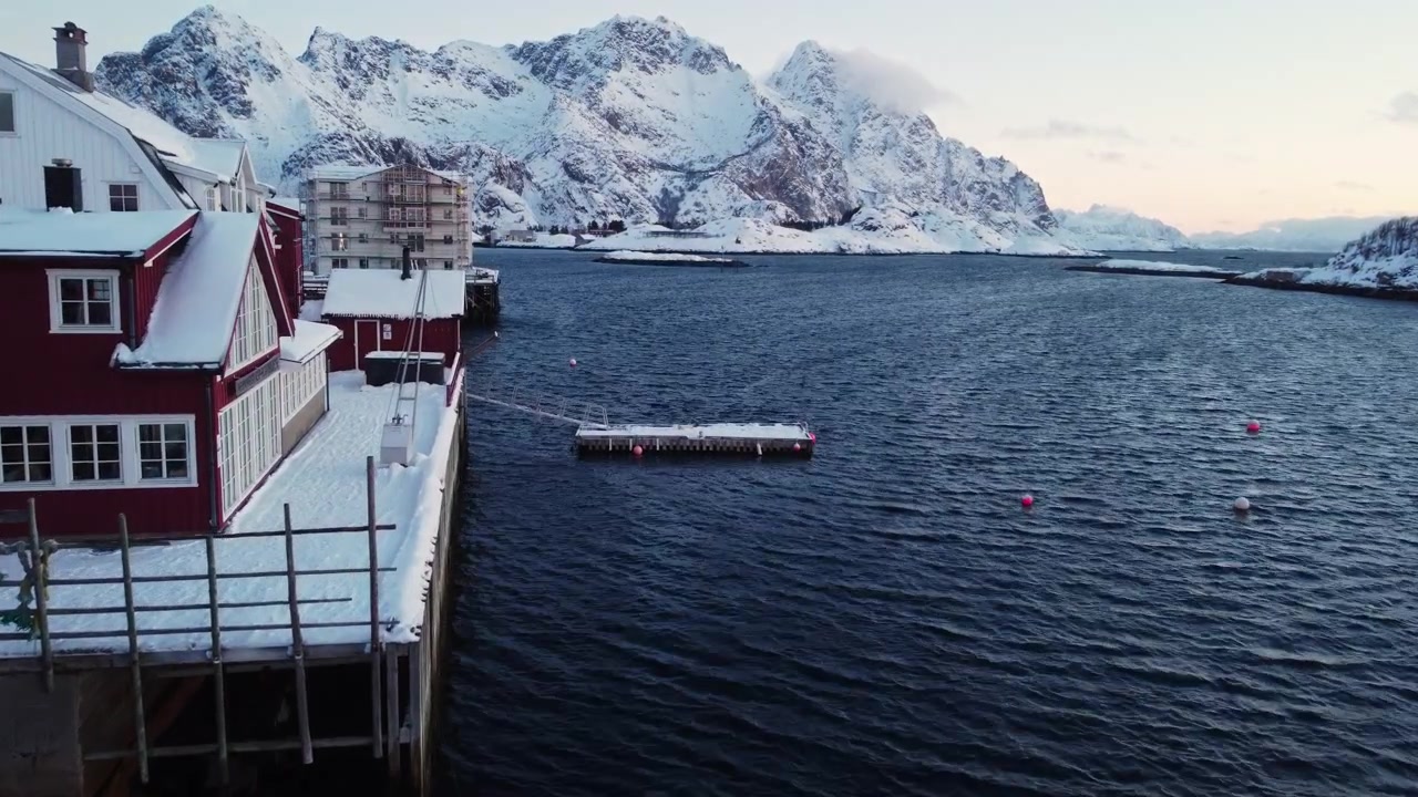 Panoramisk flyfoto av Henningsvær rorbuer ved siden av havnen med snødekte fjell i bakgrunnen, Lofoten, Norge. Berømt arktisk fiskelandsby-landskap i skumringen.