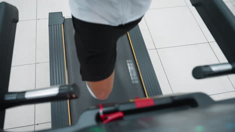 top down lower body view of athlete in white long sleeve and black shorts running on treadmill in modern gym, focus on motion of legs and treadmill belt, blurred gym floor tiles around