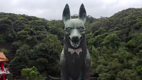 cinematic aerial backwards shot showing gigantic dog statue beside asian temple in taiwan surrounded by green forest trees in the mountains